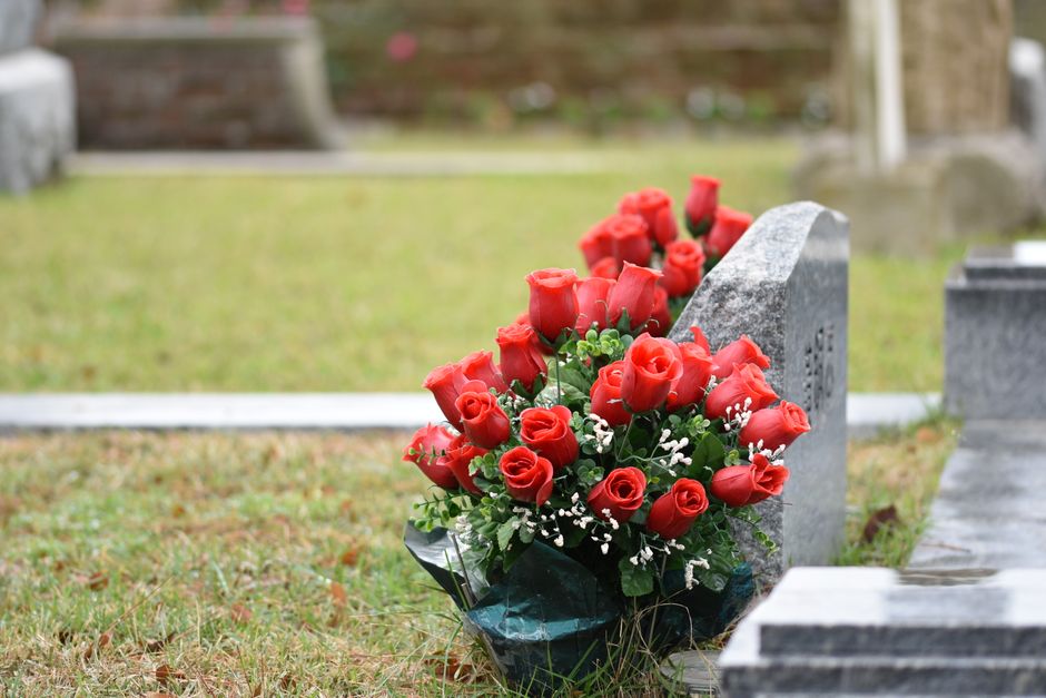 A floral arrangement of fresh red roses with tightly closed petals, interspersed with small white baby's breath and green foliage, placed on the ground against a grey stone grave marker in a cemetery 