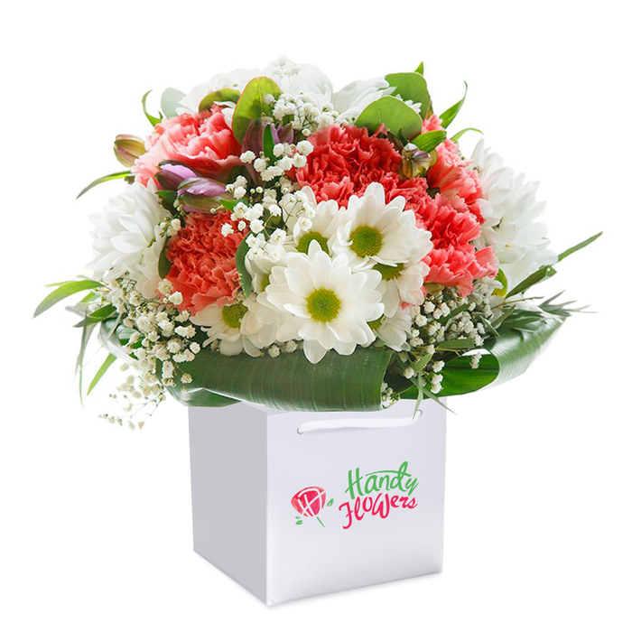 Presented against a crisp, seamless white, this awe-inspiring bouquet is nestled neatly within a square white gift bag-its clean lines and ribbon handles reflecting the understated sophistication often appreciated in Hanwell's leafy family streets. The arrangement springs to life with a rich collection of coral and fiery red carnations, each petal intricately ruffled and velvety to the eye. They are deftly complemented by snowy daisies bursting with yellow-green eyes, their brightness punctuating the bouquet's core with a joyful energy reminiscent of spring afternoons by Elthorne Park. Wispy clouds of baby's breath filter amongst the primary flowers, infusing an airy grace and highlighting the bouquet's soft-spoken innocence. Around the edges, a mantle of glossy, deep green foliage supports and frames the blossoms, while on one side, tentative purple-tipped buds bring a hush of mystery and possibility. The overall effect is one of fresh abundance-a composition that feels simultaneously structured and relaxed, suggesting a local florist's careful stewardship of every stem. The white gift bag, with its sturdy ribbon handles, promises effortless delivery to any doorstep throughout Hanwell, whether celebrating a nearby Ealing Hospital milestone or simply sending a loving thought to a neighbour by Church Road. The vivid floral colours and lush arrangement stand out crisply, inviting both sender and recipient to experience a sense of festivity, comfort, and thoughtful connection-all captured and cherished through the artistry of local floristry.
